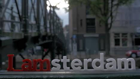 Vista-De-La-Pequeña-Figura-Plástica-De-La-Escultura-De-Letras-De-Iamsterdam-En-El-Puente-Contra-El-Paisaje-Urbano-Borroso-De-Amsterdam,-Países-Bajos