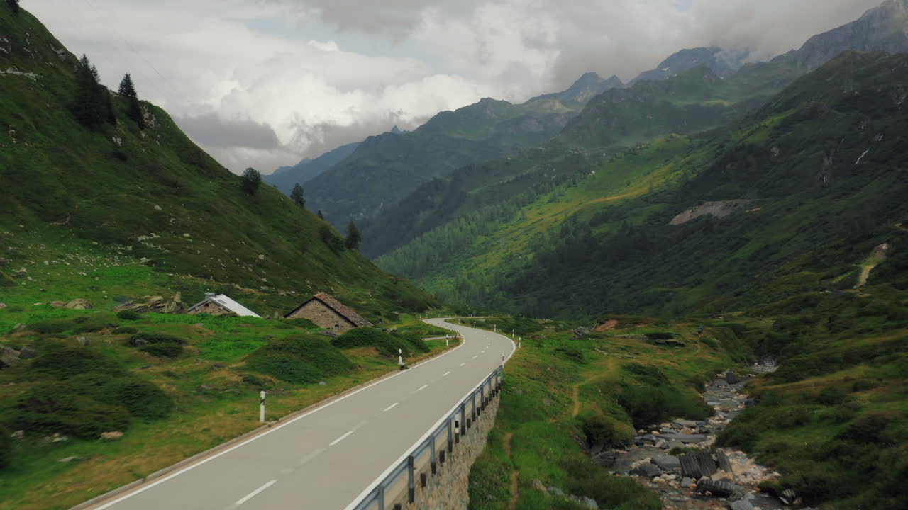 Fast wide aerial push in above epic mountain road in green valley with Alpine huts, stream and cloud-covered peaks in Switzerland. Low flying drone shot above scenic road in The Alps on a cloudy day.