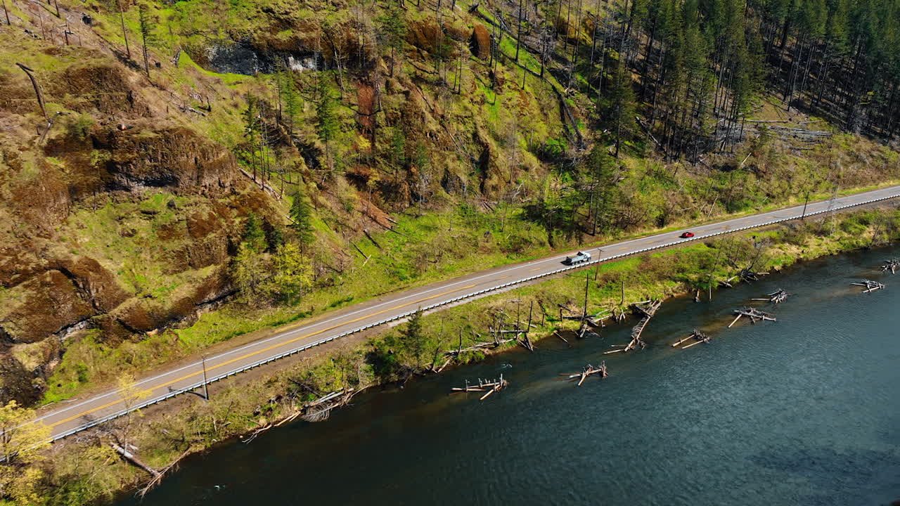 Truck follows red automobile by the mountain road near the river. Drone footage catching up with the cars moving by the wild nature.