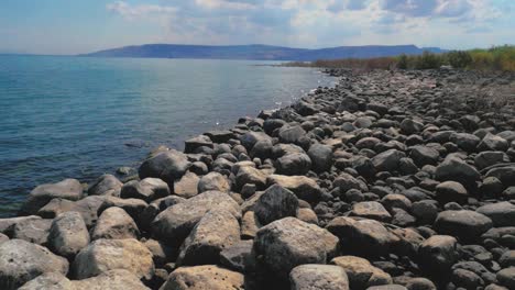 low aerial, rocky gray boulder shoreline on calm blue lake, slow motion