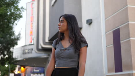 a hispanic woman standing outside a cinema movie theater building with a neon sign on a downtown urban city street