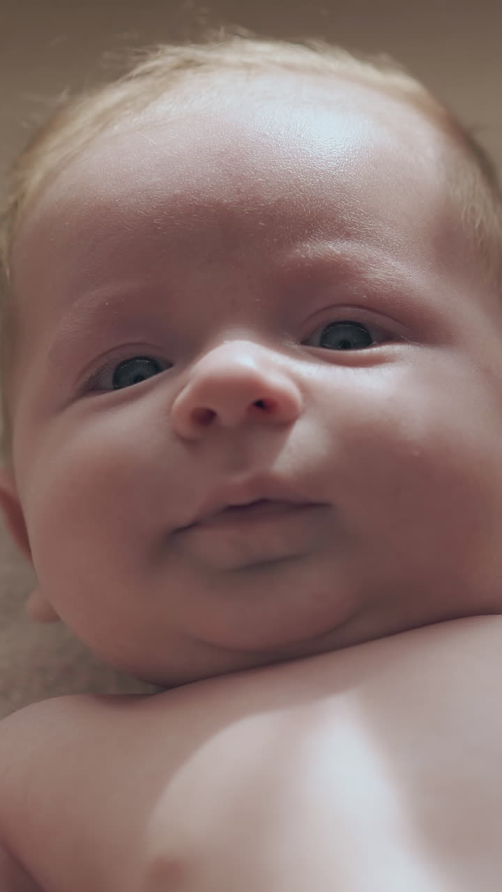 adorable newborn boy with plump cheeks and big blue eyes lies moves body and looks at camera in bed extreme close view