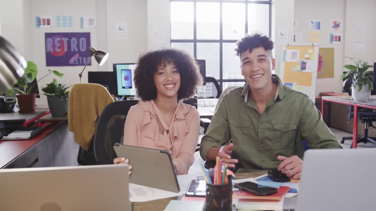 Two happy diverse creative designers in discussion at tablet in casual office, slow motion