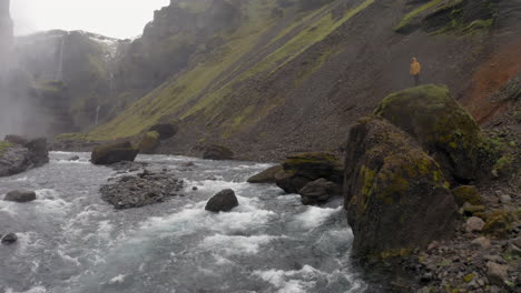 Drone-low-altitude-reveal-shot-of-one-person-with-yellow-jacket,-standing-on-a-rock-on-river-bank