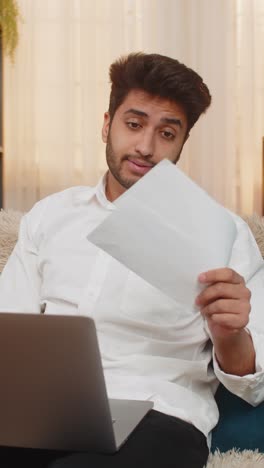 indian man suffering headache massaging temples stressed looking at laptop with scattered documents