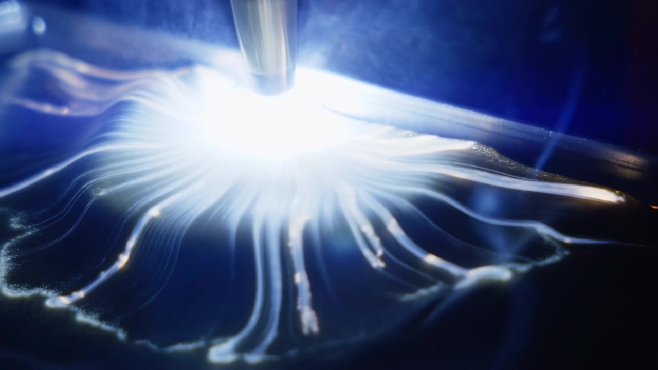 Close-up view shows intense white glow radiating outward from contact point as thin branching lines extend across dark material, with blue tones, reflective texture, and changing light patterns.