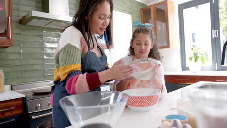 Happy-biracial-mother-and-daughter-preparing-dough-and-smiling-in-sunny-kitchen