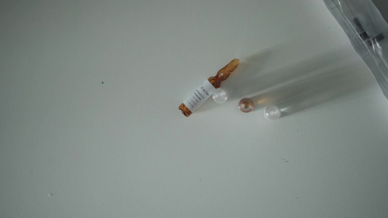 Ampolla medica e siringa sul tavolo, fiala ambrata sterile etichettata, tappo di plastica e ago nelle vicinanze, bancone clinico con illuminazione soffusa, paramedico che prepara il farmaco mentre l'infermiera verifica la dose