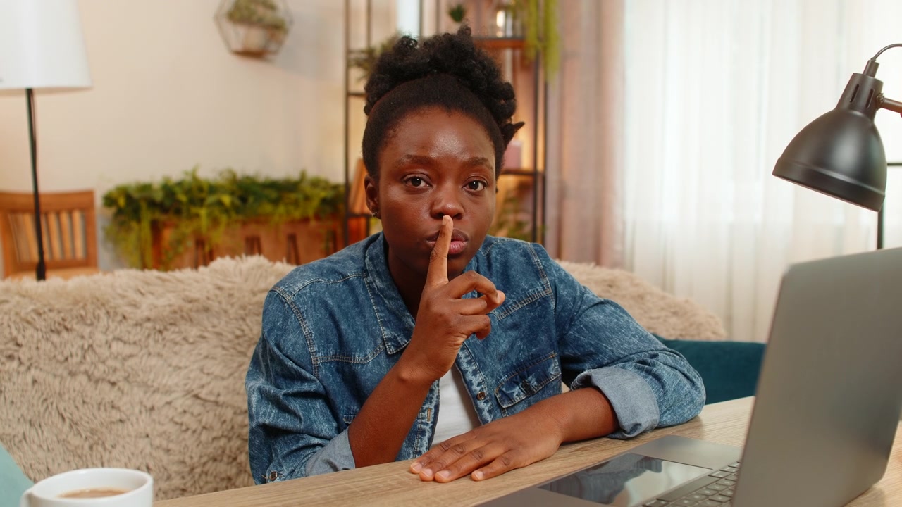 African american young woman shows silence secret gesture with finger to lips hinting secret at home