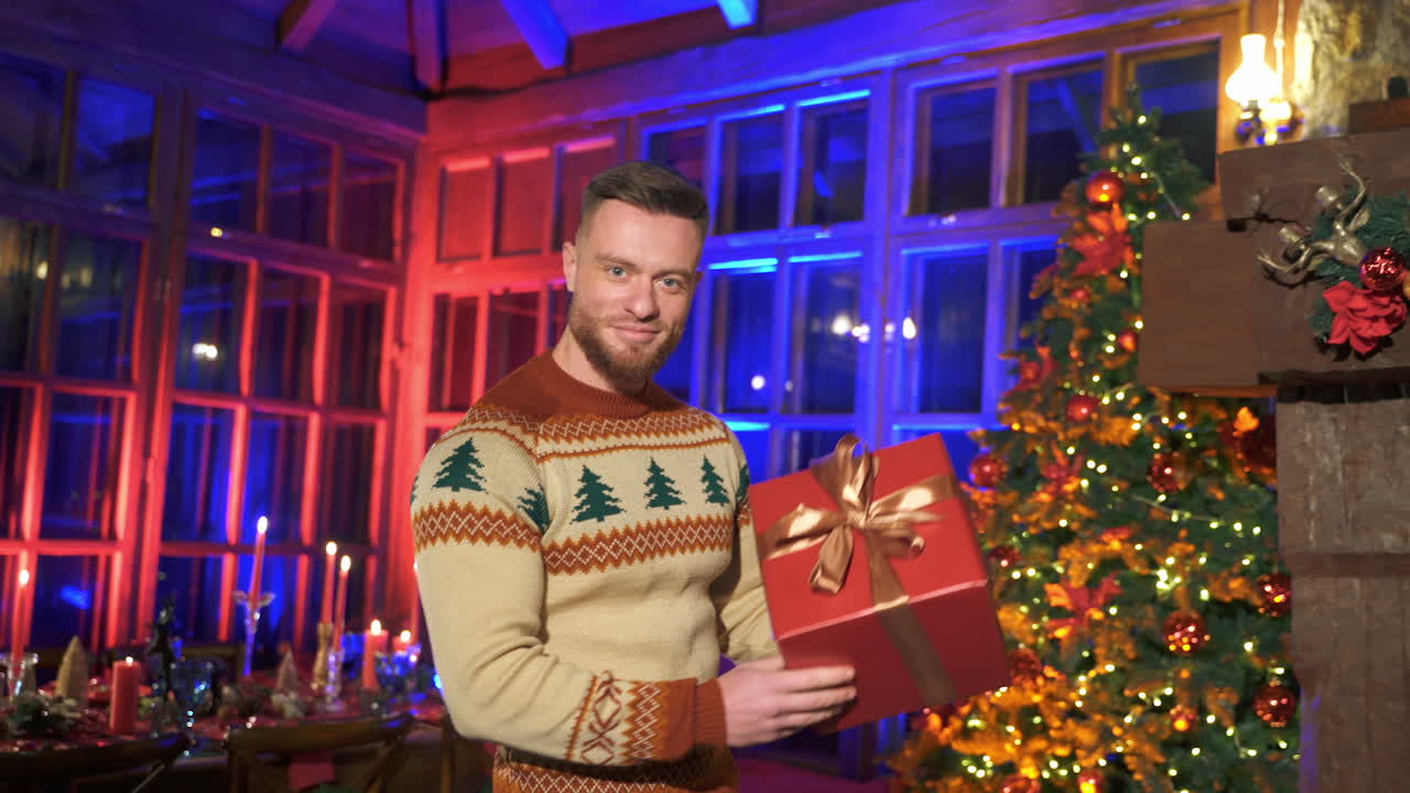 Smiling man offering new year present. Portrait of a handsome man in knitted Christmas sweater holding a box with gift on Christmas decorated room background.