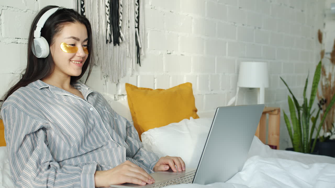 Young woman working on laptop in bed with sleep mask and headphones