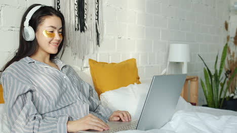 young woman working on laptop in bed with sleep mask and headphones