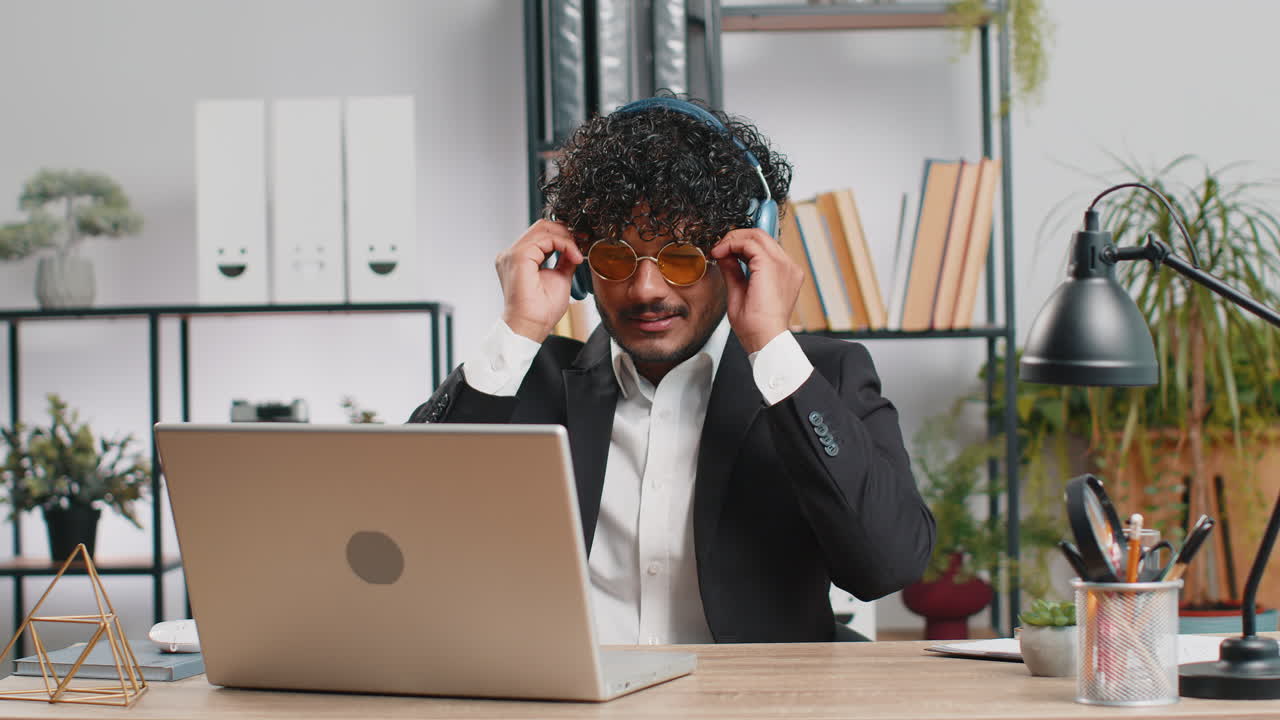 Indian businessman wearing headphones listening music dancing after working on laptop at home office