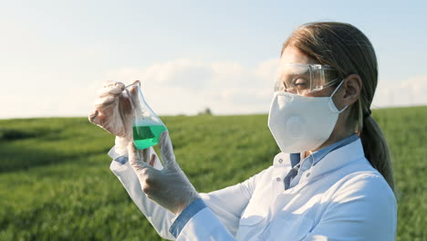 close-up view of caucasian researcher woman in white coat and goggles looking at test tube in the green field