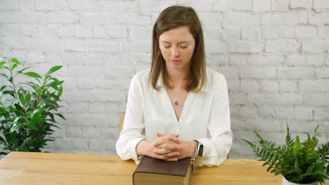 a young woman praying with a bible and fingers clasped together in prayer sitting at a table