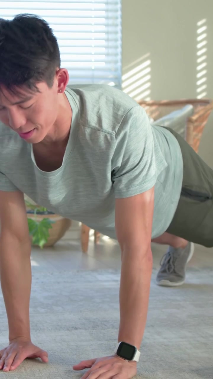Vertical video: Starting low, man doing pushups at home on rug in shorts for fitness, dumbbell near