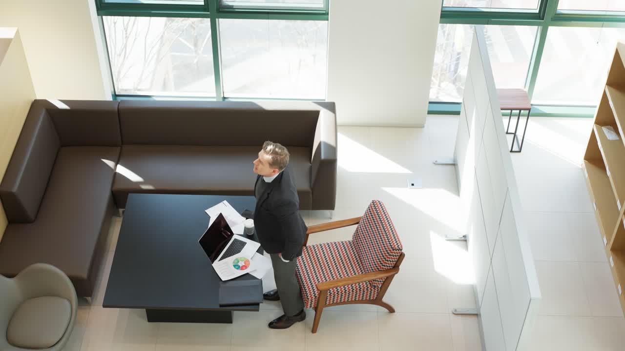 A thoughtful caucasian businessman stands up from an armchair crossing his arms near a table with a laptop and scattered paper documents - top down view