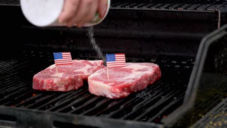 Zwei-Saftige-Rib-Eye-Steaks-Sitzen-Auf-Dem-Grill-Und-Kochen-Mit-Zwei-Winzigen-Zähnen-Der-Amerikanischen-Flagge,-Während-Salz-In-Zeitlupe-Auf-Die-Steaks-Gegossen-Wird