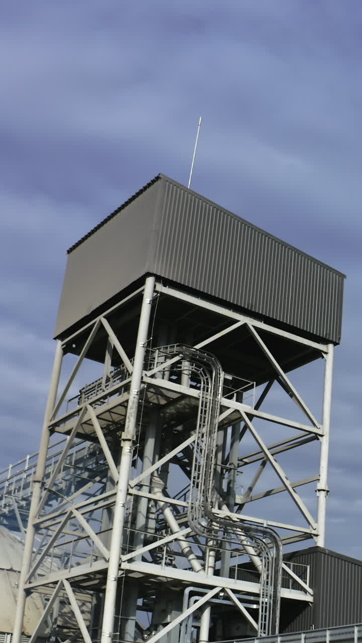 High metal construction standing next to the elevator tanks. Looking at the tops of modern granary buildings at the backdrop of grey cloudy sky. Vertical video