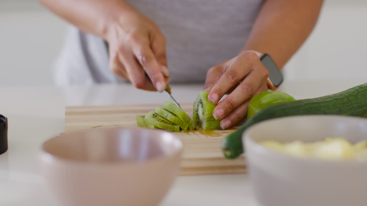 Donna che tiene fermo un kiwi dimezzato, affettando fette sottili con un coltello da cucina su un tagliere di legno per uno spuntino.