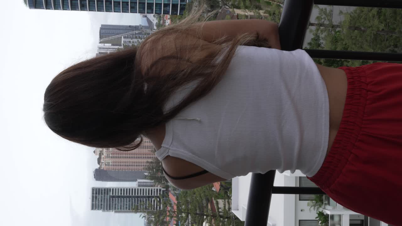 mujer morena sexy en pijama mirando desde el balcón del apartamento hacia una ciudad moderna