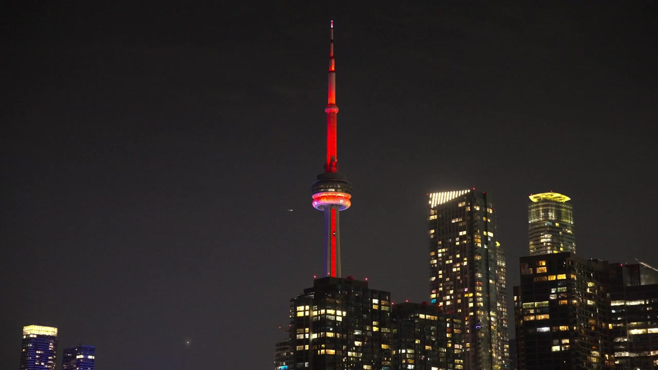 La CN Tower illuminata di notte si erge sopra gli edifici del centro di Toronto mentre la telecamera si muove lentamente da sinistra a destra.
