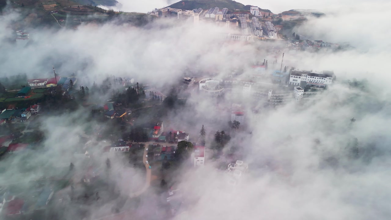Aerial drone footage of a mountain town in Sapa surrounded by dense fog and low clouds with buildings and winding streets appearing through mist in northern Vietnam highlands