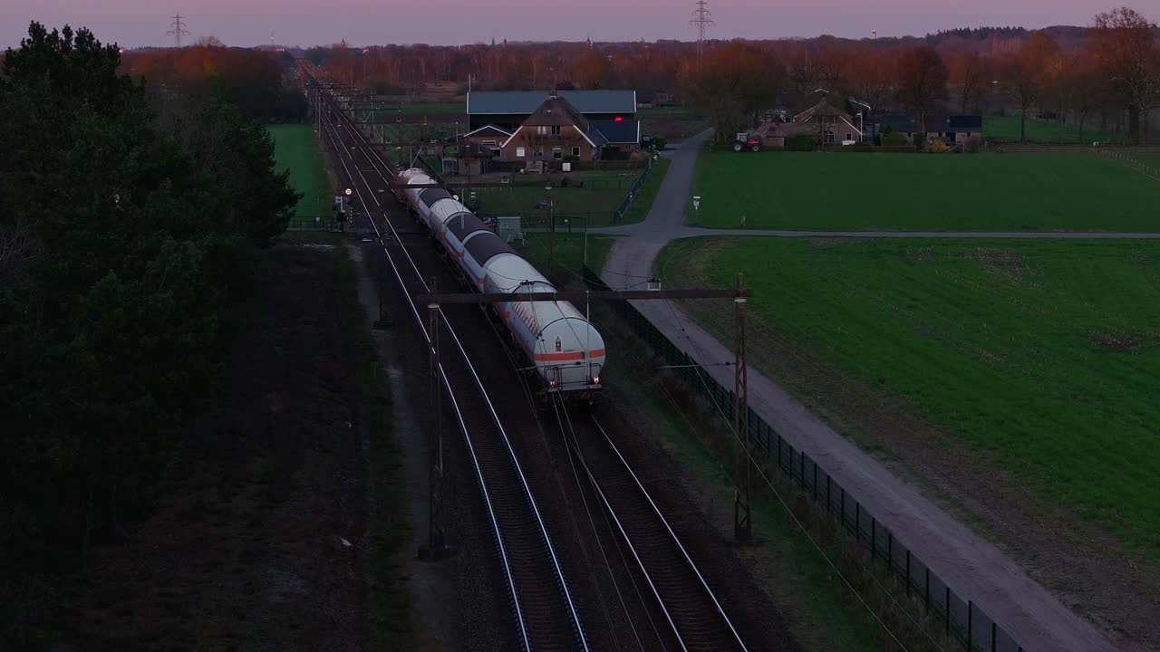 Long cargo train with cylindrical tanker wagons moves through a rural Dutch village at dusk, flanked by farmhouses and fields under a purple-orange sky, drone static shot, real time