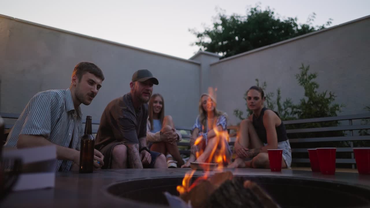 A confident blond guy lights a fire in a barbecue while relaxing and partying in the courtyard of a country house during twilight