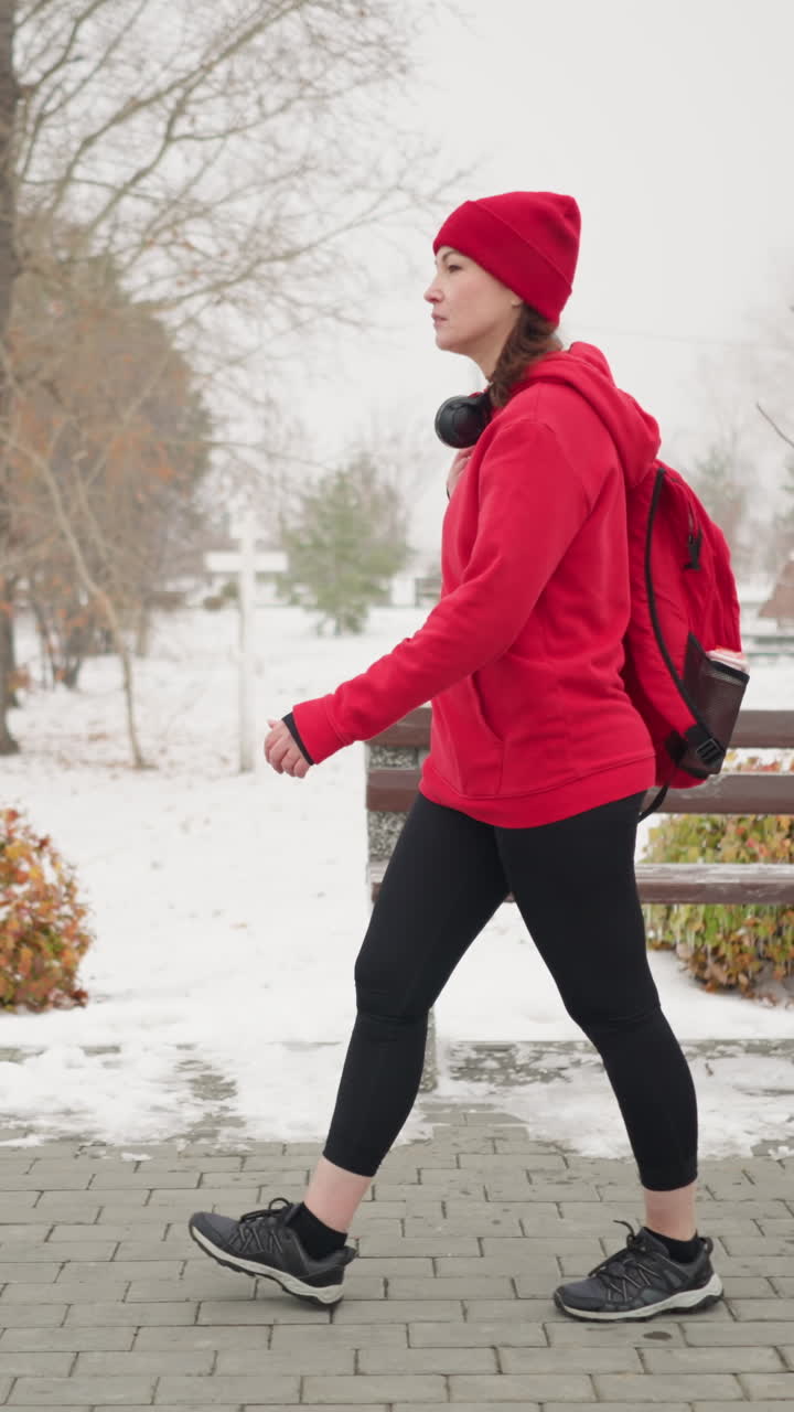 Side view of woman with bag over shoulder walking along interlocked pathway surrounded by snowy park scenery, benches, colorful bushes, and light poles in foggy atmosphere during winter