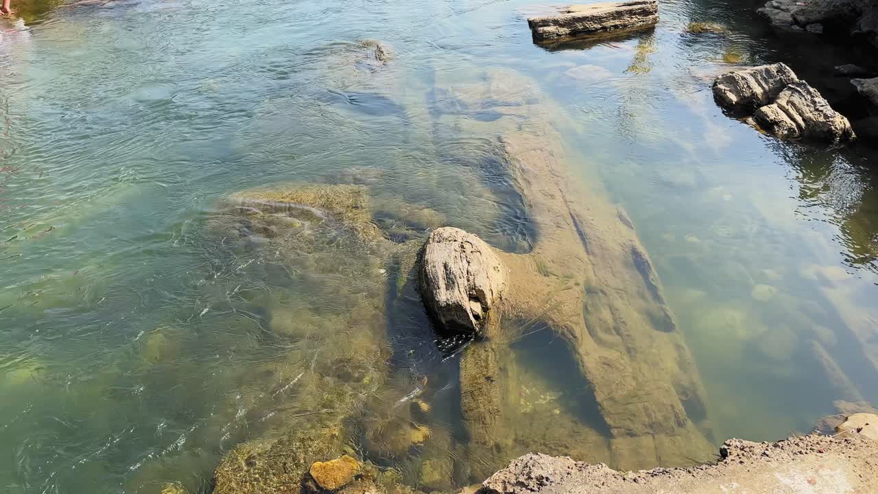 Vista dall'alto di acqua di fiume limpida che scorre dolcemente su rocce sommerse, rivelando trame di pietre lisce, bassa profondità e motivi naturali sotto la luce brillante del giorno.