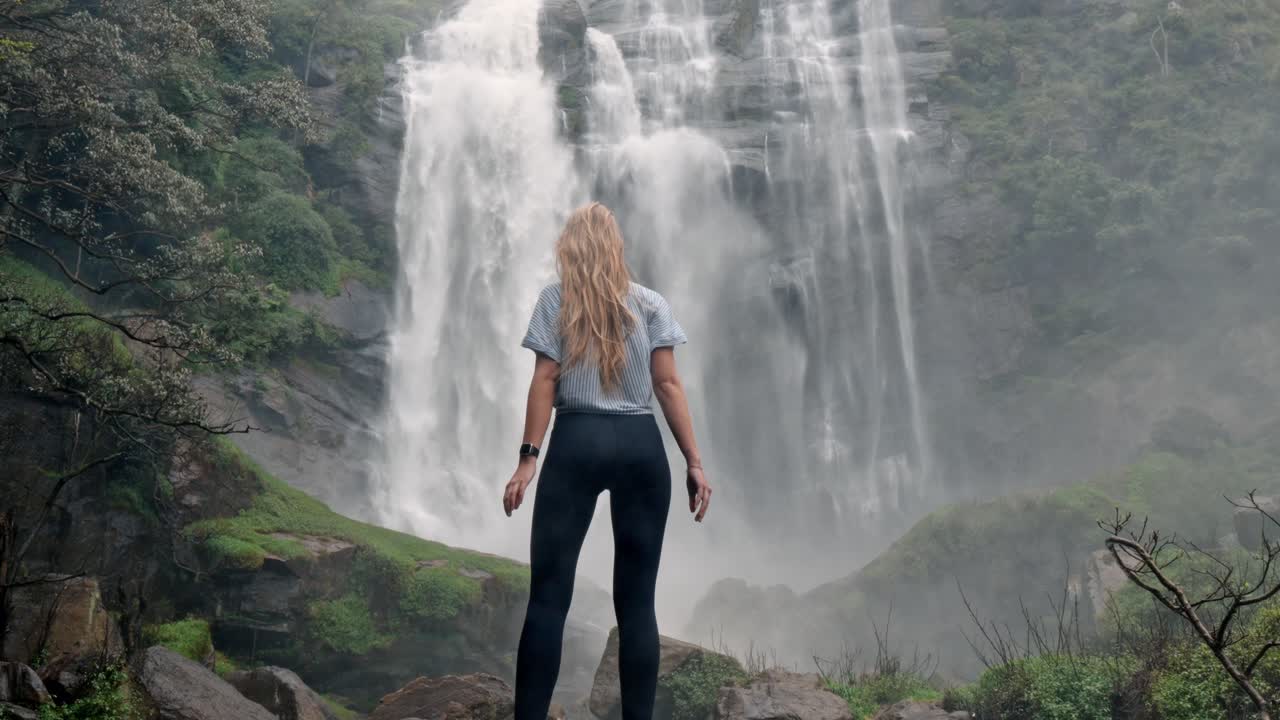 A woman stands in awe before the roaring Bomburu Ella Waterfalls, surrounded by mist, jungle, and rocky cliffs in the lush highlands of Nuwara Eliya, Sri Lanka.