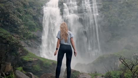 A woman stands in awe before the roaring Bomburu Ella Waterfalls, surrounded by mist, jungle, and rocky cliffs in the lush highlands of Nuwara Eliya, Sri Lanka.