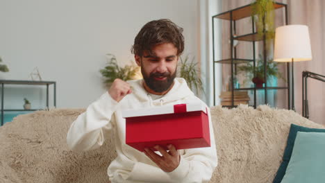 happy young indian man holding birthday gift box with ribbon congratulating sitting on sofa at home