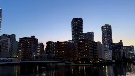 Skyline-Der-Stadt-In-Der-Abenddämmerung-Mit-Beleuchteten-Gebäuden,-Die-Sich-In-Einem-Ruhigen-Fluss-Spiegeln