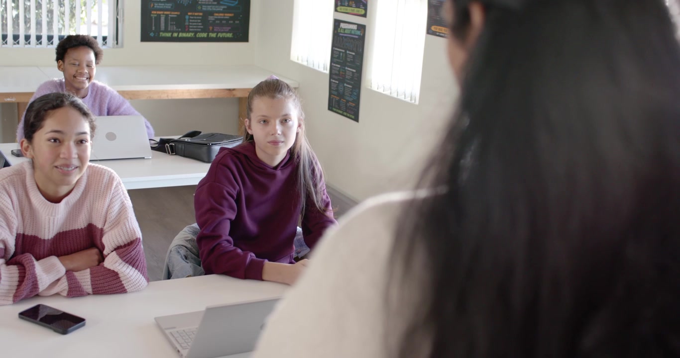 Diverse female teacher leading lesson, gesturing to 3 teen girls around laptop in class, copy space