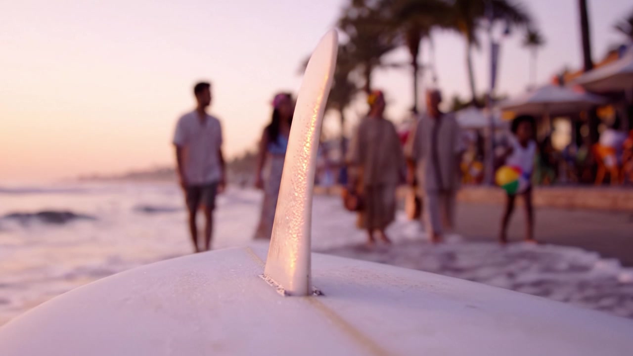 A Serene Evening at the Beach: Surfers and Beachgoers Stroll Along the Shoreline as Waves Gently Crash Alongside a Surfboard in Warm Sunset Glow