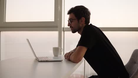 relaxed caucasian man in stylish glasses using laptop, having video call. cheerful smiling man in black clothes sitting on chair having a friendly video call. happy man enjoying video calling at big, bright house. close up