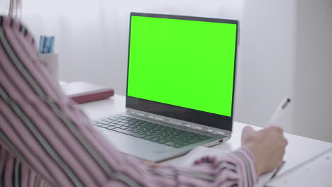 una mujer joven está aprendiendo a escribir en un cuaderno y viendo la pantalla verde en una computadora portátil para la tecnología chroma key