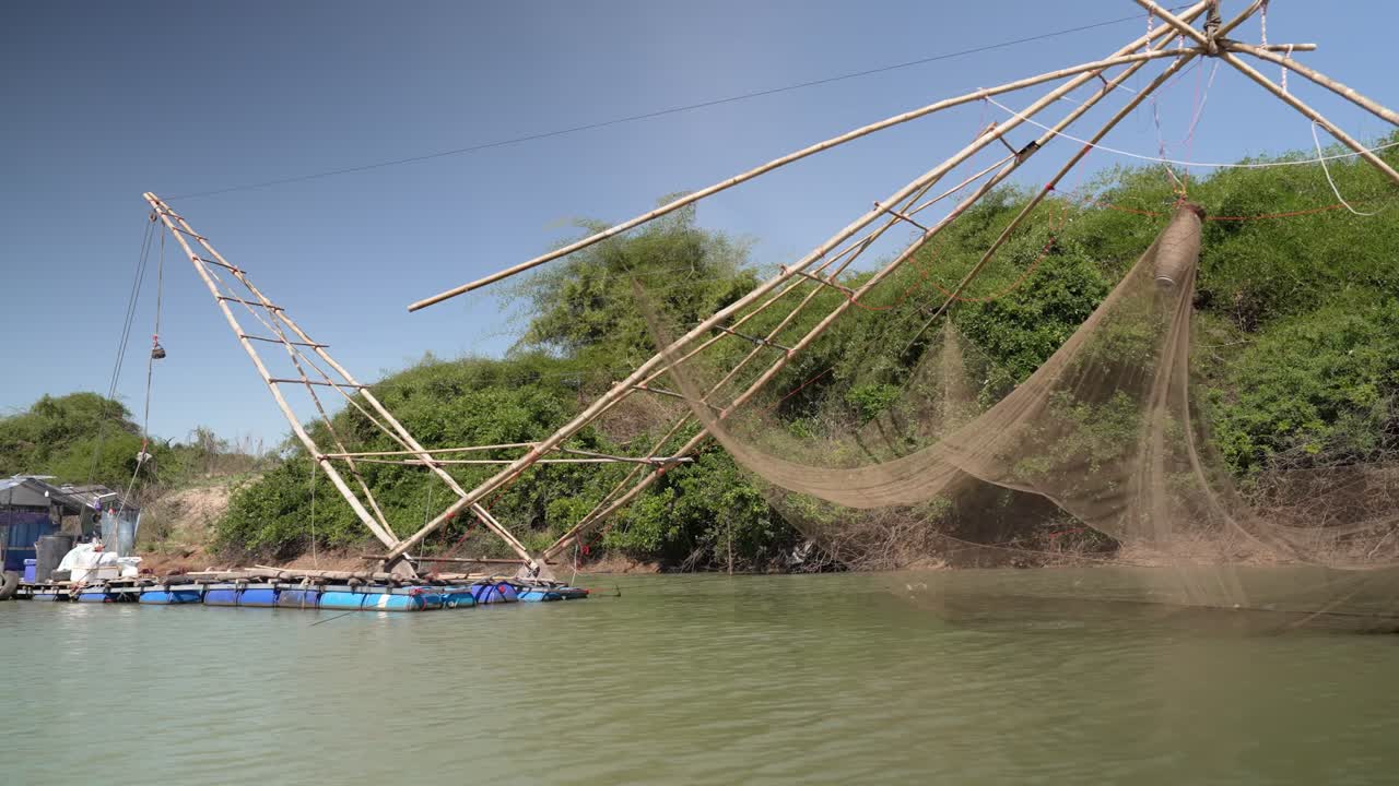 Wide shot of a bamboo lift-net rig on a small riverside raft, calm river water and bright sky creating a serene, traditional fishing scene