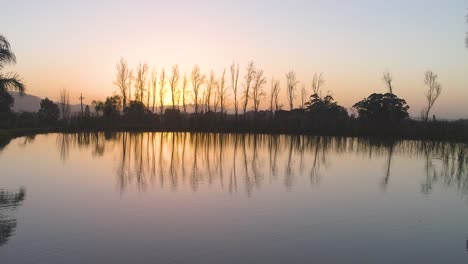 Drone-flies-low-over-dam-showing-silhouette-of-surrounding-trees-reflecting-off-surface-during-golden-sunset