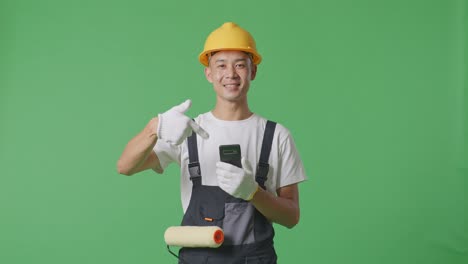 asian man painter wearing safety helmet smiling and pointing to smartphone in his hand while standing in the green screen background studio