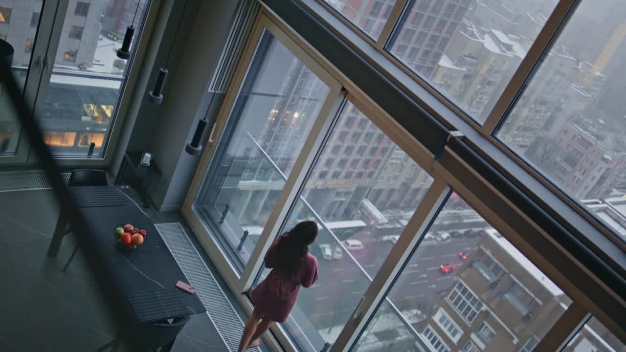 Mulher em casa pensando no futuro, olhando pela janela panorâmica em apartamento, em plano de ângulo alto