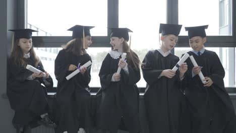 grupo de felices estudiantes de preescolar multirraciales con birrete y bata. están sentados y sosteniendo diplomas.