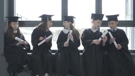 group of happy multiracial preschool students in mortarboard and gown. they are sitting and holding diplomas.