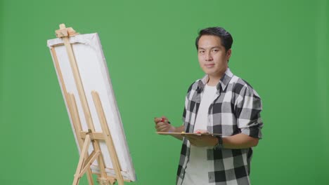 asian male artist smiling to camera while painting on canvas by oil paints and brush in the green screen studio