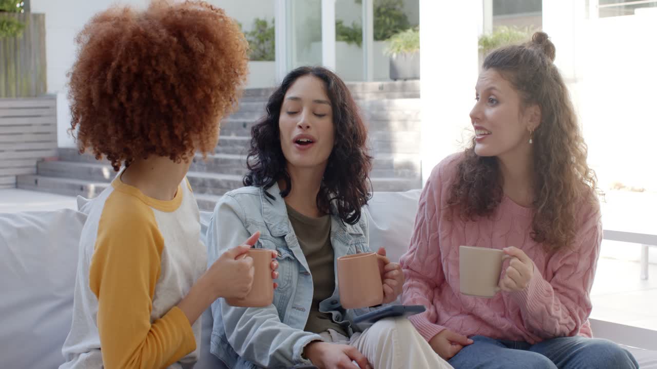 Amiche di etnie diverse che tengono in mano tazze di ceramica e sorridono, reagendo a notizie sorprendenti in un patio soleggiato