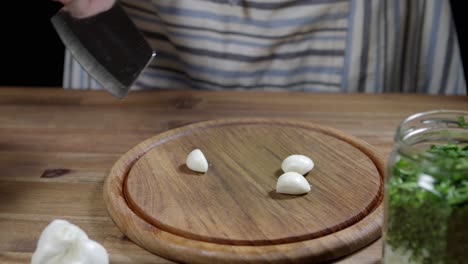 chef cutting stems of three cloves of garlic, uses kitchen ax knife on round wooden board