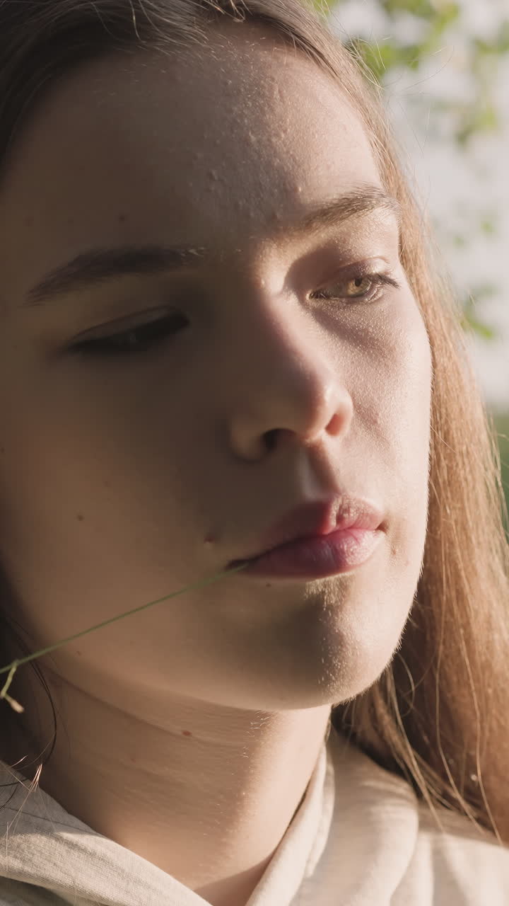 mujer con hoja de hierba en la boca se apoya en el tronco del árbol. luz del sol en el lado de la cara. la joven tiene problemas psicológicos sintiendo ansiedad. aislamiento en la naturaleza al atardecer