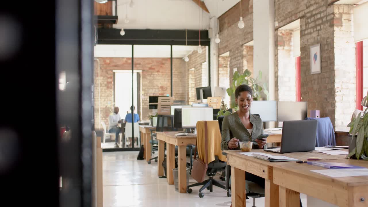 Happy african american casual businesswoman using smartphone and having lunch in office, slow motion
